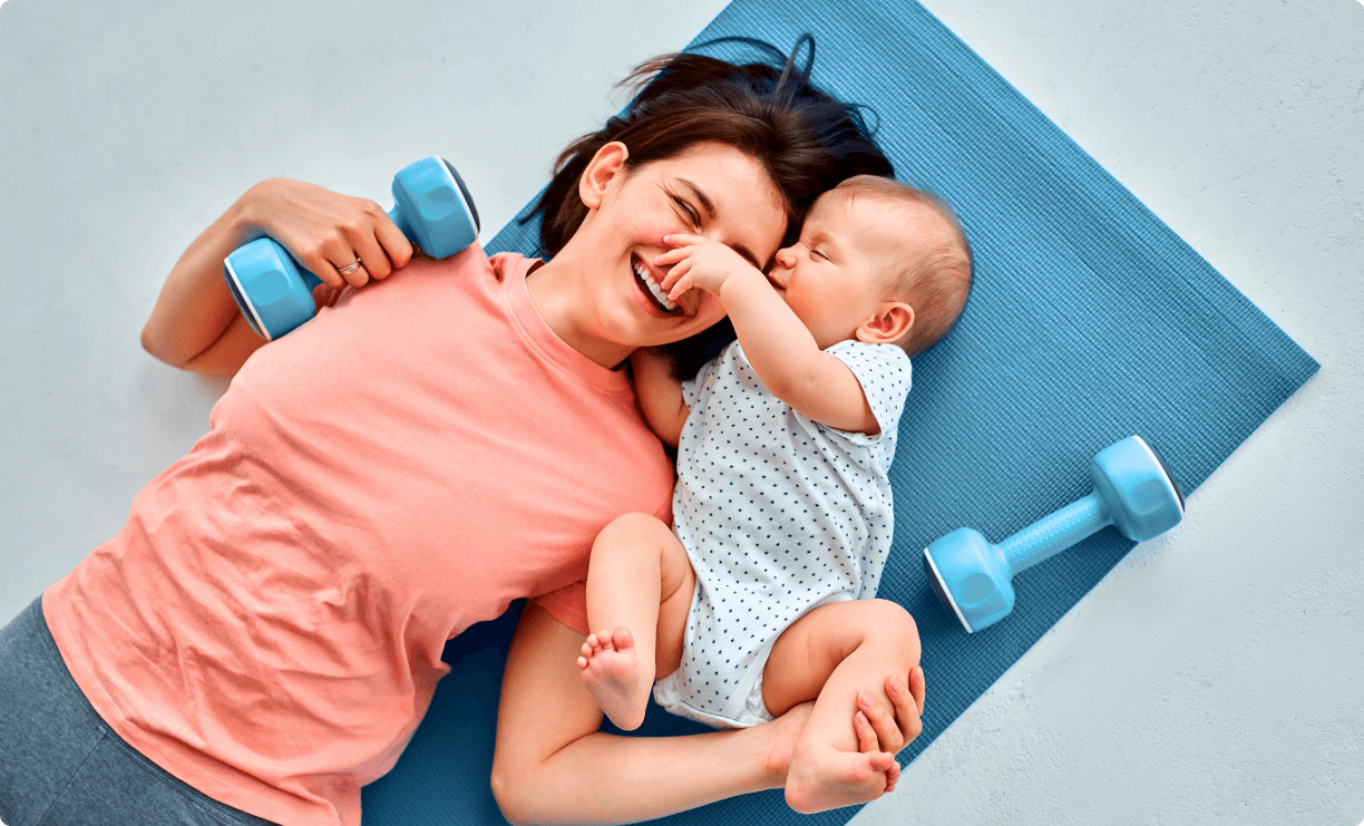 Top view of a healthy mother with baby boy lying on a yoga matt and playing with dumbbells.