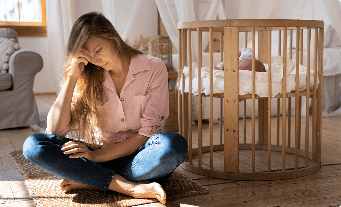 A tired woman sits on the floor, head in hand, while a baby lies in a round wooden crib behind her.