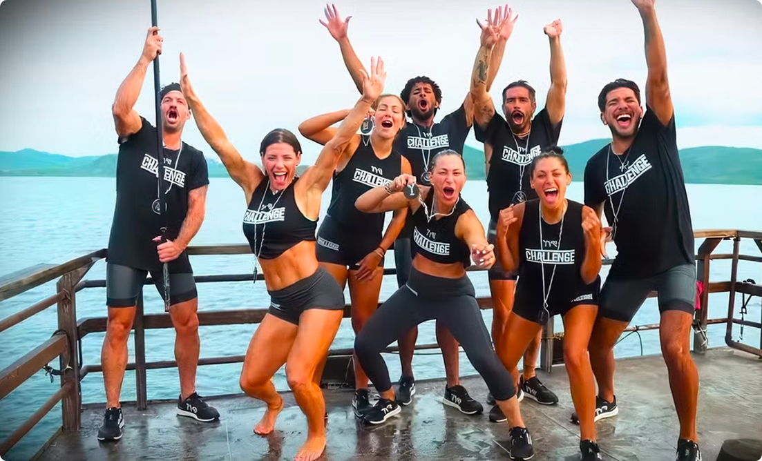 A group of athletic contestants from The Challenge TV show celebrating on a dock, dressed in matching black outfits, expressing excitement and victory near a scenic waterfront.